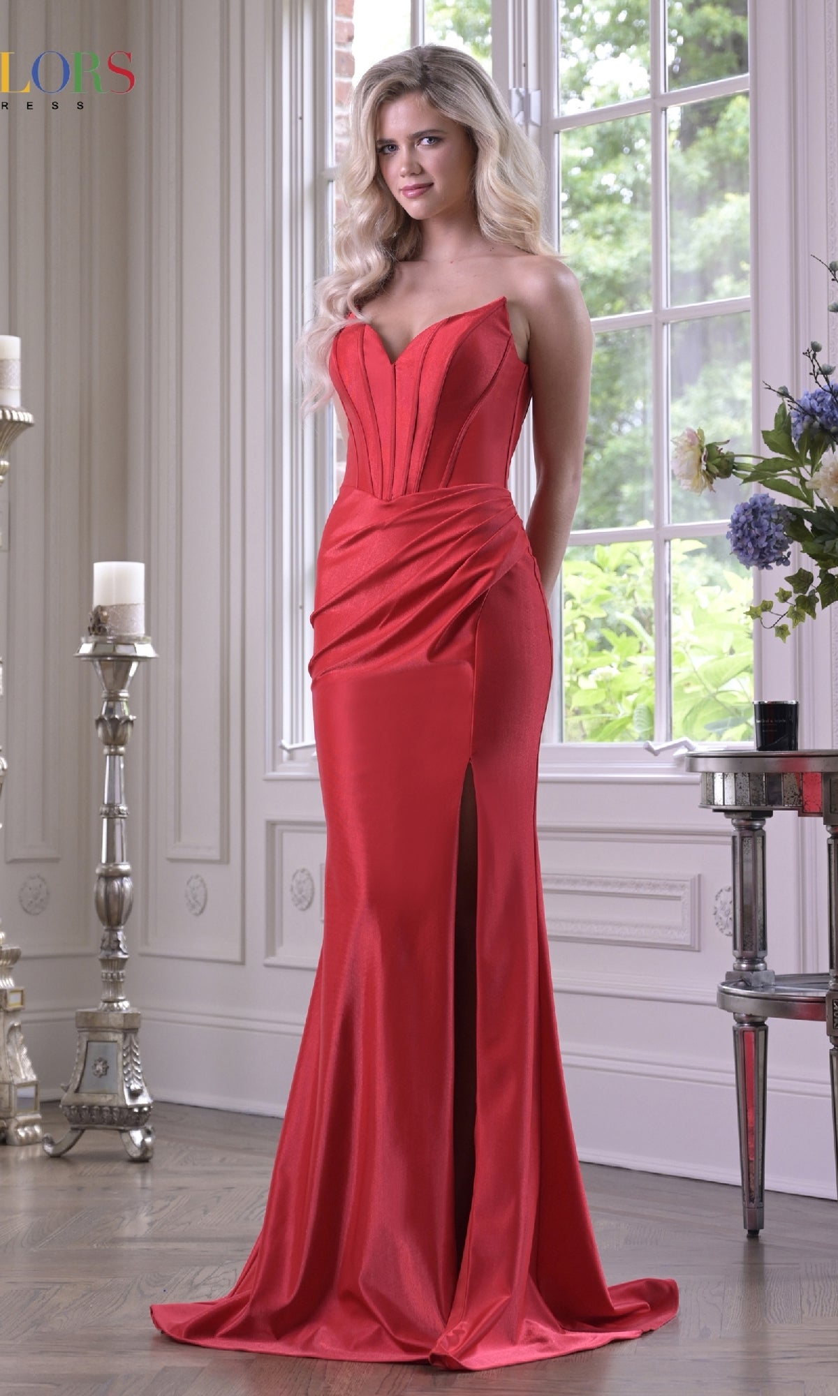 A woman in the red Strapless Long Corset Prom Dress: Colors Gown 3839 stands indoors by a large window, classic decor around her and greenery visible outside, the dress featuring a thigh-high slit.